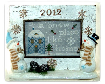 May include: A white wooden frame with a cross-stitch design inside. The design features a house with a blue and white pattern, a snow-covered tree, and the text "Snow place like home". Two snowman figurines are on either side of the frame.