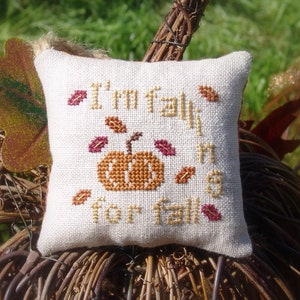 May include: A white cushion with the text "I'm fall-ing for fall" and a small orange pumpkin with a green stem embroidered on it. The cushion is decorated with small red and orange leaves.