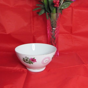 May include: A white porcelain bowl with a floral design sits on a red surface. The bowl has a gold rim and a pink and green floral pattern. A vase with a bouquet of white, pink, and yellow flowers is in the background.