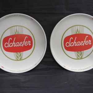 May include: Two white metal serving trays with a gold and red design. The trays feature the text "Schaefer" in a white cursive font on a red oval. The text "Schaefer Beer" is written in a smaller font around the edge of the tray.