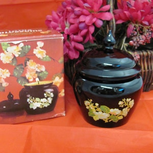 May include: A black ceramic jar with a lid and floral design. The jar is filled with a candle and has a label that reads "BLOSSOMS" on the front. The jar is sitting on a red surface.