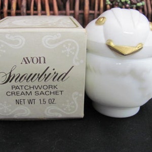 May include: A white ceramic snowbird-shaped cream sachet with gold accents. The box reads "Avon Snowbird Patchwork Cream Sachet Net Wt 1.5 oz."