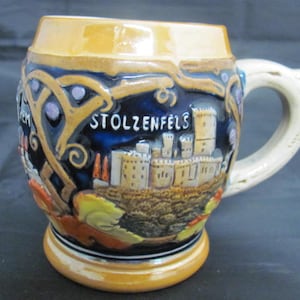 May include: A ceramic beer stein with a brown, blue, and white design. The stein features a castle with the text "Stolzenfels" on the front.
