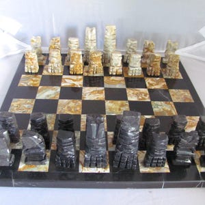 May include: A complete chess set with black and tan stone pieces on a black and tan checkered board. The chess pieces are carved in a unique, abstract style. The board is made of a polished stone material.