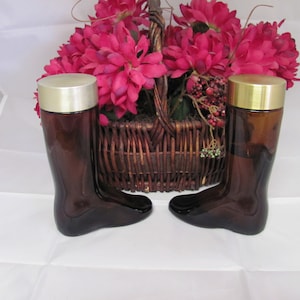 May include: Two brown glass bottles shaped like boots with gold lids. The bottles are sitting on a white surface in front of a wicker basket filled with pink flowers.
