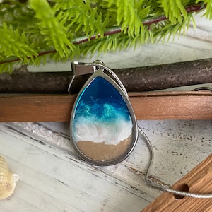 Puede incluir: Un collar con un colgante en forma de l&aacute;grima que contiene una imagen de resina de una escena oce&aacute;nica. El colgante representa agua azul, espuma blanca y arena marr&oacute;n.