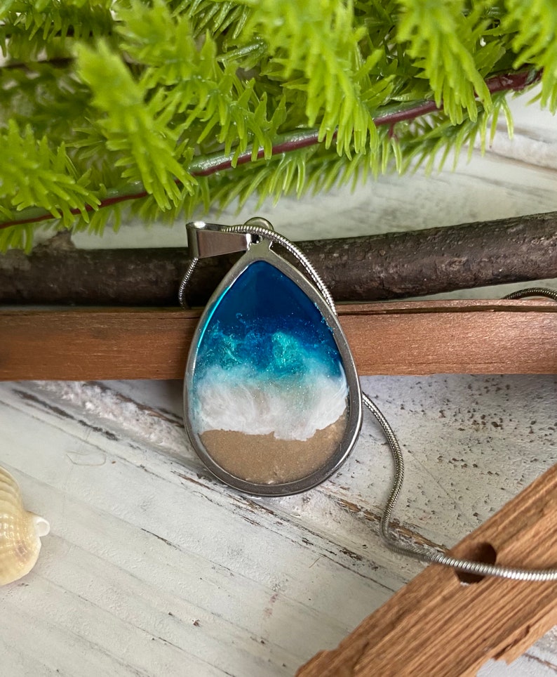Puede incluir: Un collar con un colgante en forma de l&aacute;grima que contiene una imagen de resina de una escena oce&aacute;nica. El colgante representa agua azul, espuma blanca y arena marr&oacute;n.