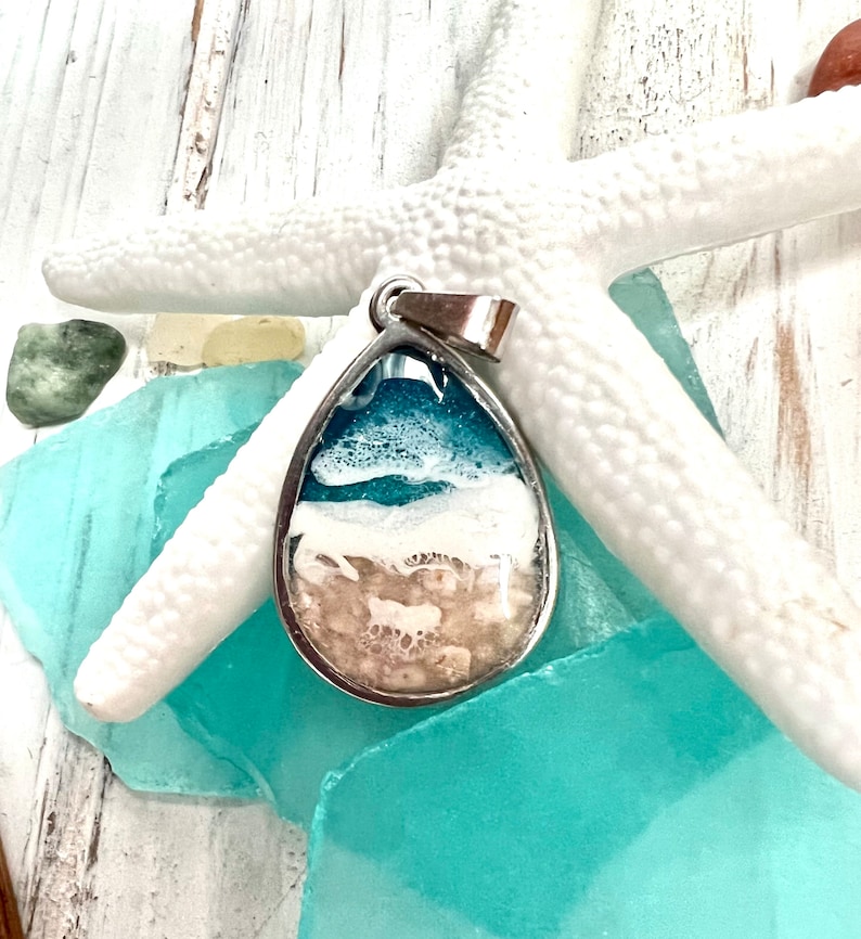 Puede incluir: Un colgante en forma de l&aacute;grima con una imagen de resina de una escena de playa. El colgante est&aacute; hecho de plata y tiene una argolla de plata. La imagen de resina muestra un oc&eacute;ano azul, olas blancas y una playa de arena.