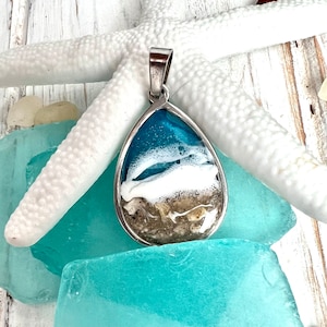 Puede incluir: Un collar con un colgante en forma de l&aacute;grima con una incrustaci&oacute;n de resina que representa una escena de playa. La escena incluye agua azul, espuma blanca y arena marr&oacute;n. El colgante es plateado y tiene un asa para sujetar una cadena.