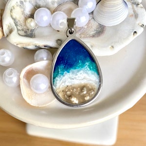 Puede incluir: Un collar con un colgante en forma de l&aacute;grima que representa una escena de oc&eacute;ano en resina. El colgante presenta un oc&eacute;ano azul con olas blancas que rompen en una playa de arena. El colgante est&aacute; rodeado de perlas blancas y conchas.