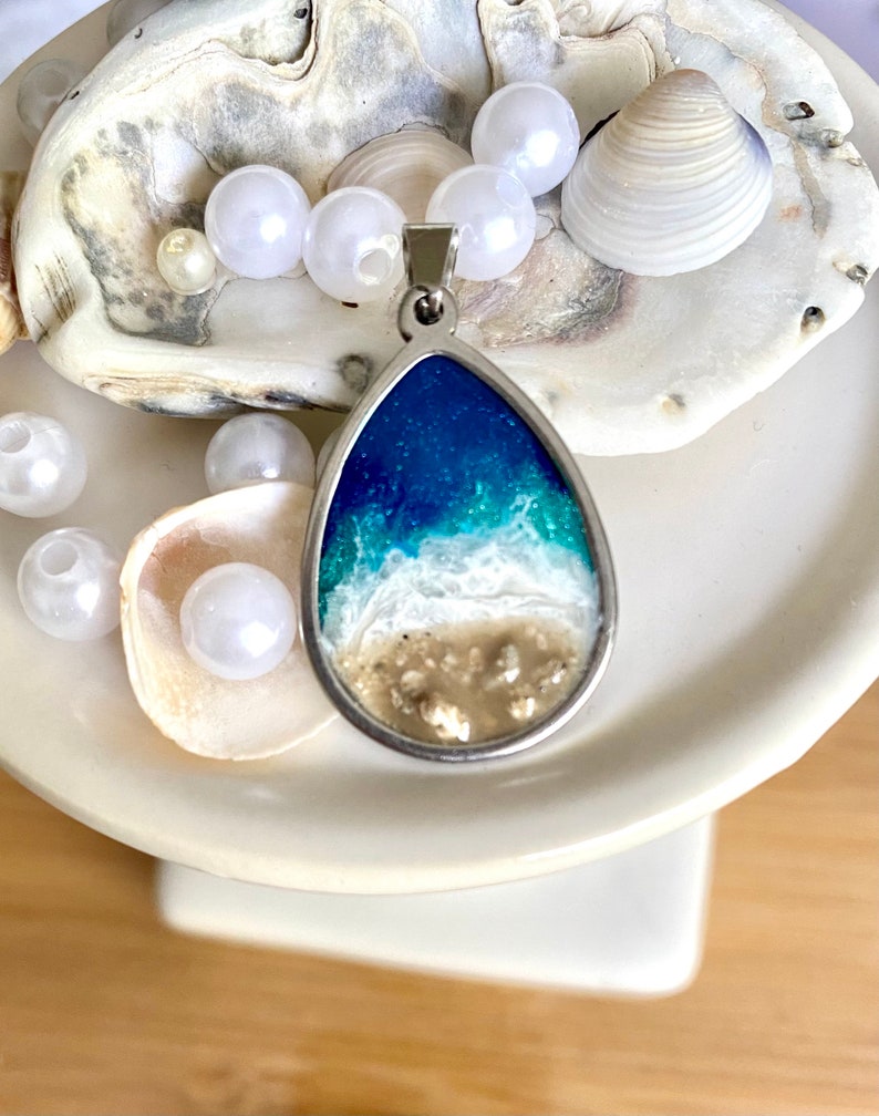 Puede incluir: Un collar con un colgante en forma de l&aacute;grima que representa una escena de oc&eacute;ano en resina. El colgante presenta un oc&eacute;ano azul con olas blancas que rompen en una playa de arena. El colgante est&aacute; rodeado de perlas blancas y conchas.