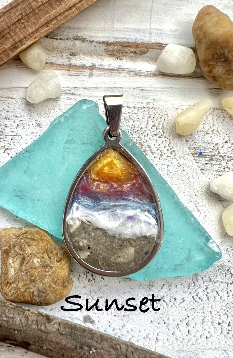 Puede incluir: Un colgante de plata con una escena de puesta de sol en su interior. La escena presenta una puesta de sol amarilla, naranja, roja y morada sobre una ola blanca y arena gris. El colgante est&aacute; sobre una superficie azul claro con la palabra "Sunset" escrita debajo.