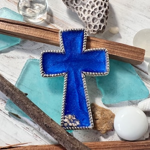 May include: A silver-toned cross pendant with a vibrant blue center. The cross has a detailed border and a textured surface. The pendant is displayed with natural elements like sea glass and coral, suggesting a coastal or religious theme.