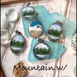 Puede incluir: Cinco colgantes de plata con diferentes escenas de monta&ntilde;a y lago. Cada colgante presenta un paisaje diferente con monta&ntilde;as verdes, agua azul y nubes blancas. Los colgantes se muestran sobre un fondo azul y blanco.