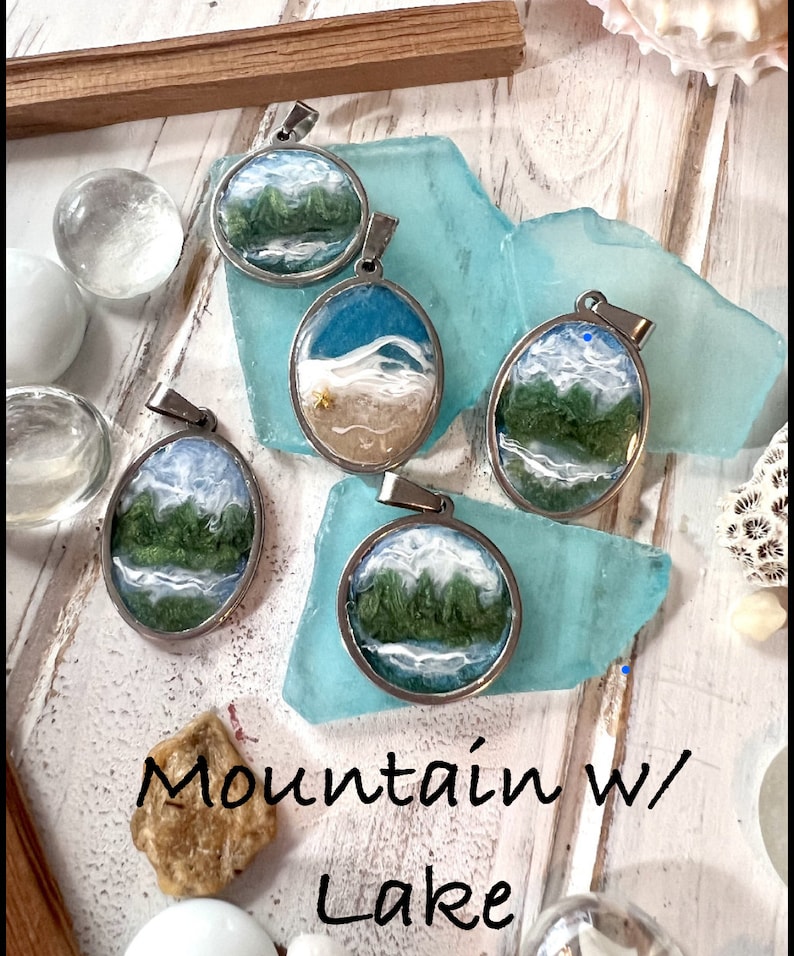Puede incluir: Cinco colgantes de plata con diferentes escenas de monta&ntilde;a y lago. Cada colgante presenta un paisaje diferente con monta&ntilde;as verdes, agua azul y nubes blancas. Los colgantes se muestran sobre un fondo azul y blanco.