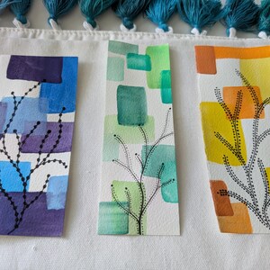 Puede incluir: Tres marcapáginas de acuarela con diseños abstractos. Cada marcapáginas presenta una combinación de colores única: azul y morado, verde y amarillo y naranja. El arte lineal negro representa un patrón similar a una rama.