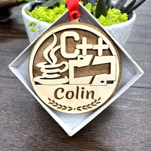 May include: A wooden ornament with a red ribbon, featuring a stylized C++ logo with a coffee cup and the name "Colin" inscribed below.