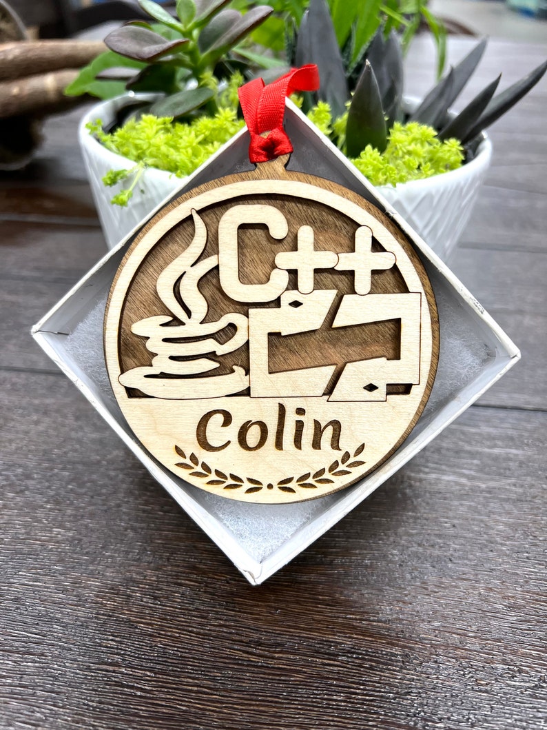 May include: A wooden ornament with a red ribbon, featuring a stylized C++ logo with a coffee cup and the name "Colin" inscribed below.