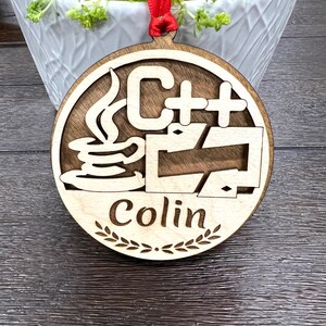 May include: A round wooden ornament with a red ribbon. The ornament features a stylized image of a coffee cup with steam rising from it, the text "C++", and a stylized arrow. The ornament also has the name "Colin" written on it.