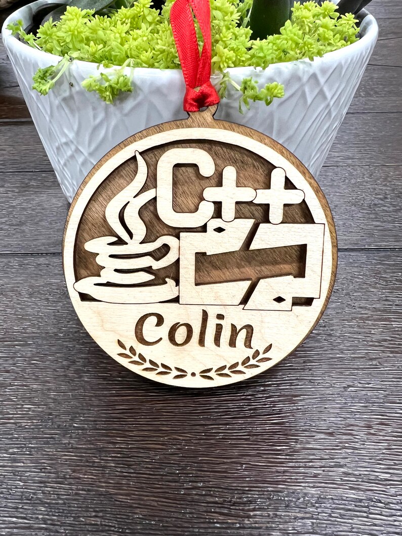 May include: A round wooden ornament with a red ribbon. The ornament features a stylized image of a coffee cup with steam rising from it, the text "C++", and a stylized arrow. The ornament also has the name "Colin" written on it.