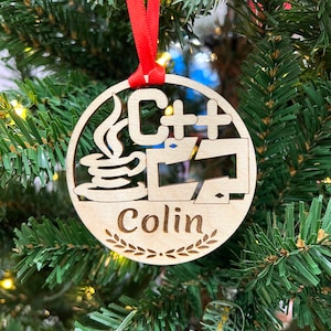 May include: A wooden Christmas ornament with a red ribbon. The ornament features a stylized image of a coffee cup with steam, the text "C++", and a computer monitor. The ornament also includes the name "Colin" with a laurel wreath.