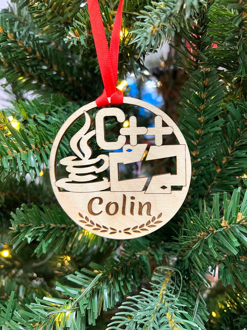 May include: A wooden Christmas ornament with a red ribbon. The ornament features a stylized image of a coffee cup with steam, the text "C++", and a computer monitor. The ornament also includes the name "Colin" with a laurel wreath.