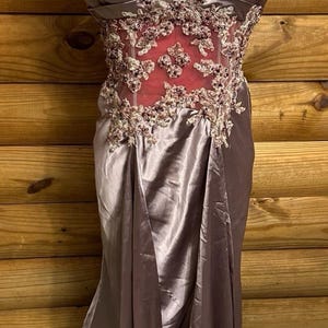 Könnte beinhalten: Ein formelles, bodenlanges Kleid in gedämpftem Mauve, das auf einer Schneiderpuppe präsentiert wird. Das Oberteil hat aufwendige Blumenstickereien und einen Sweetheart-Ausschnitt. Das Kleid hat einen fließenden Rock und zarte Träger.