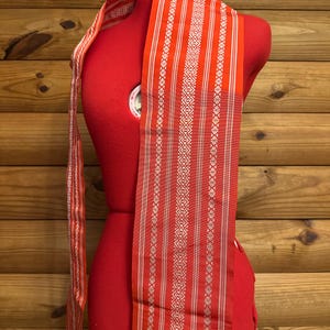 Puede incluir: Un textil naranja y blanco estampado está drapeado sobre un maniquí rojo. El textil presenta rayas verticales y intrincados diseños geométricos blancos. La tela probablemente se utiliza para ropa o fines decorativos.