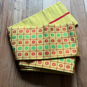 May include: A folded yellow fabric sash featuring a grid pattern formed by thin red lines. Each square in the grid contains a small embroidered flower in either red or green. A single horizontal red stripe is visible on the plain yellow section.