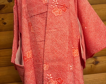 May include: A red kimono with a floral pattern. The kimono is made of a textured fabric and features white and orange flower designs. The garment is displayed on a mannequin, showcasing its shape and style.