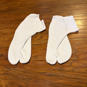 May include: A pair of white tabi socks featuring a split-toe design that separates the big toe from the other toes. The socks have a low-cut ankle height and include a small metal hook-and-loop closure detail at the top of the ankle opening.