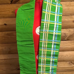Puede incluir: Una faja de tela verde, plisada por un lado y estampada por el otro. El diseño de cuadros presenta rayas blancas, amarillas y azules sobre un fondo verde. La faja está drapeada sobre un maniquí rojo.