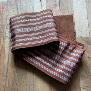 May include: A folded length of fabric in a rich copper-brown hue, featuring horizontal stripes in white and silver. The textile displays a repeating geometric pattern woven into the bands, creating a textured appearance across the surface.