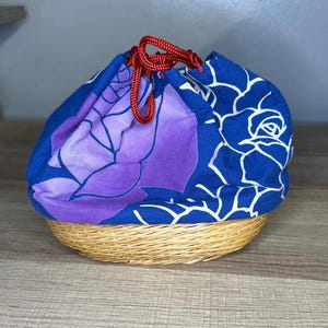 May include: A round woven basket with a blue and purple floral drawstring bag. The bag features a large rose design in shades of blue and purple. The drawstring is red. The basket is made of natural-colored woven material.