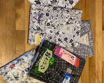 May include: A collection of five folded yukata garments in clear plastic packaging. The items feature various patterns including blue and white floral prints, a dark grey plaid design, and a blue and white grid pattern, each labeled with size M tags.