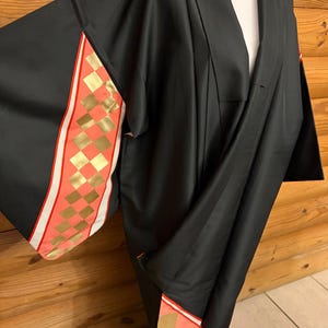 Könnte beinhalten: Ein schwarzer Kimono mit weiten Ärmeln und einem dekorativen Einsatz mit korallenrosa und goldfarbenen Karomustern. Das Kleidungsstück hat einen V-Ausschnitt und lange, fließende Ärmel, wobei sich das Karomuster am unteren Saum wiederholt.