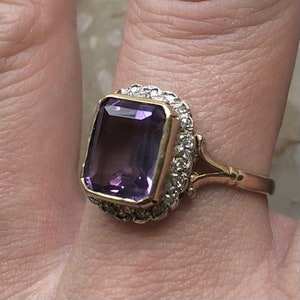 Könnte beinhalten: Ein goldener Ring mit einem großen, rechteckigen, violetten Amethyst-Edelstein, der von einem Heiligenschein aus kleinen, klaren Diamanten umgeben ist.