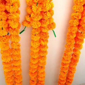 Puede incluir: Tres hileras de guirnaldas de flores de cal&eacute;ndula de color naranja brillante, cada una con varios racimos de flores, colgando de una pared blanca.