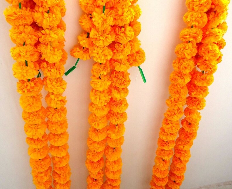 Puede incluir: Tres hileras de guirnaldas de flores de cal&eacute;ndula de color naranja brillante, cada una con varios racimos de flores, colgando de una pared blanca.