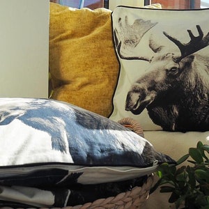 May include: Two decorative throw pillows with a black and white moose print and a yellow pillow. The pillows are on a white sofa with a green plant in front of them.