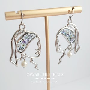 May include: Silver earrings with a silhouette of a woman's face, featuring a pressed flower design and a pearl accent. The earrings are displayed on a gold stand.
