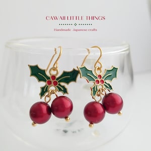 May include: Gold-tone dangle earrings featuring a holly leaf design with green enamel and red berry accents. The earrings have a red pearl-like bead hanging from each leaf. The text "CAWAII LITTLE THINGS" and "Handmade Japanese crafts" is visible.