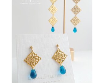 May include: Gold-colored filigree earrings with a diamond-shaped design and a teardrop-shaped blue bead. The earrings are displayed on a white card with text that reads "CAWAII LITTLE THINGS" and "Handmade Japanese crafts".