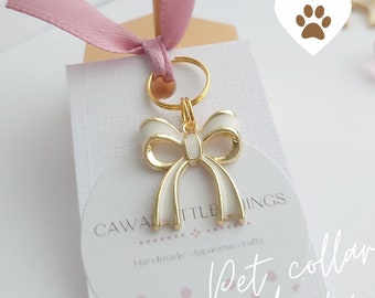 May include: White enamel bow charm with gold trim and a gold split ring. The charm is on a white card with the text "Cawaiittle Things Handmade Japanese crafts Pet collar charm".