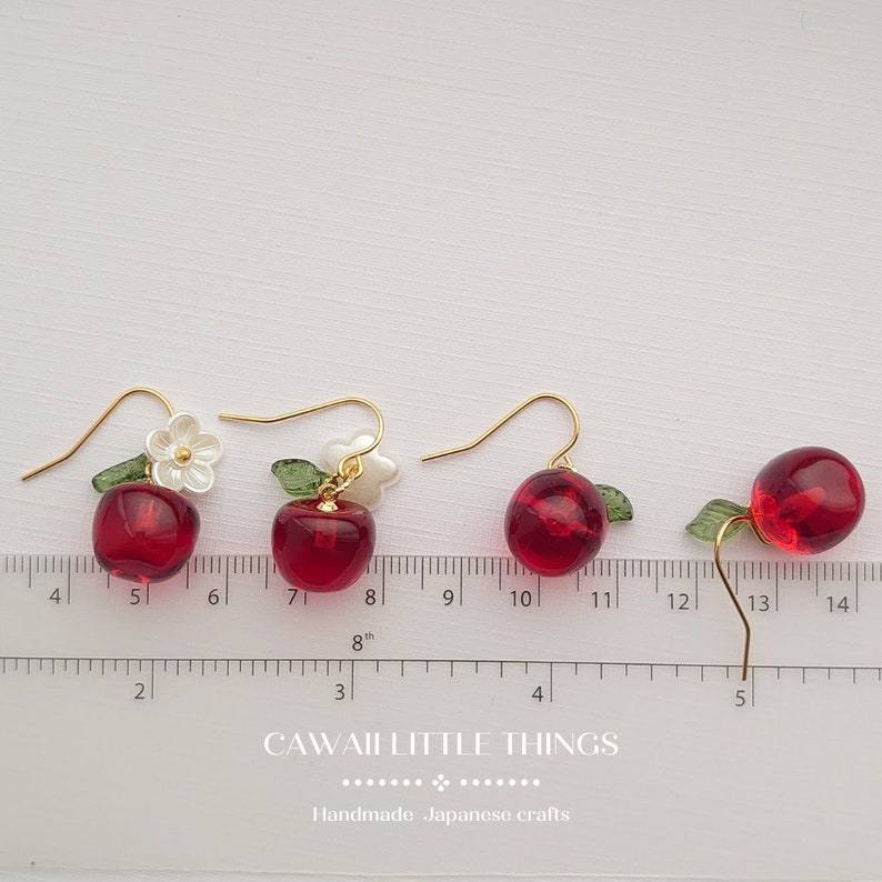 Puede incluir: Cuatro pares de pendientes con forma de cereza con ganchos dorados. Las cerezas son de un rojo transl&uacute;cido con hojas verdes. Algunos pendientes tienen detalles de flores blancas. El texto "CAWAII LITTLE THINGS" y "Handmade Japanese crafts" son visibles.