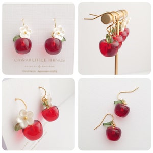 Puede incluir: Pendientes rojos con forma de manzana con ganchos dorados, detalles de flores blancas y hojas verdes. Los pendientes se muestran sobre un fondo blanco. El texto "CAWAI LITTLE THINGS" es visible.