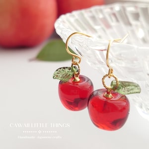 Puede incluir: Un par de pendientes artesanales de manzana de cristal rojo con ganchos dorados y detalles de hojas verdes. Los pendientes se exhiben cerca de un cuenco de cristal y una manzana roja borrosa. El texto "CAWAII LITTLE THINGS" y "Handmade Japanese crafts" es visible.