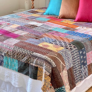 Peut inclure: Une courtepointe patchwork aux motifs et couleurs variés, notamment du bleu, du marron, du rose et de l'orange. Le lit est orné de coussins bleus, marron et roses. Une lampe blanche et un vase avec des roses rouges sont sur une table de chevet.