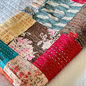 May include: A patchwork quilt made of various fabrics with floral and geometric patterns in shades of pink, red, brown, yellow, blue, and white. The quilt is folded and lying on a white surface.