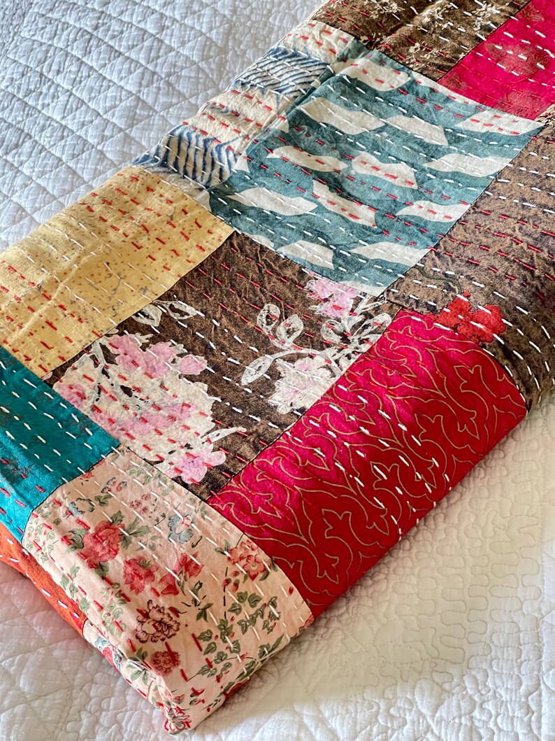 May include: A patchwork quilt made of various fabrics with floral and geometric patterns in shades of pink, red, brown, yellow, blue, and white. The quilt is folded and lying on a white surface.